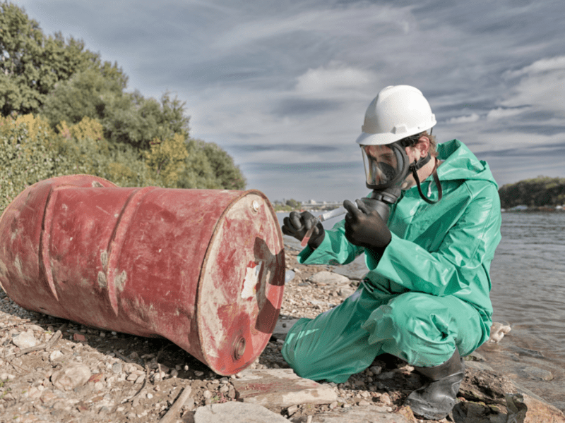 Photograph of a worker in a green hazard suit and mask inspecting a barrel along a lake side.